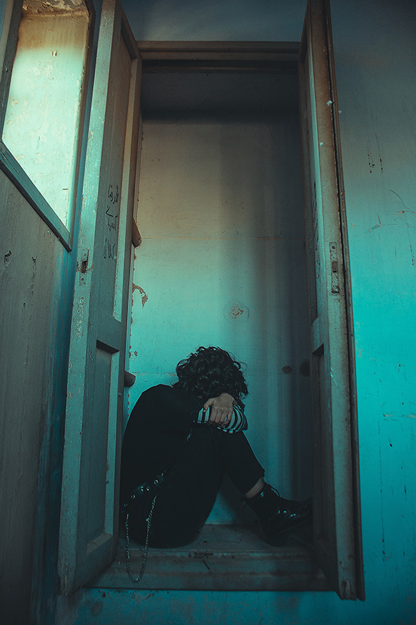 A person wearing black boots with a curly hair sitting in a doorway, looking out.