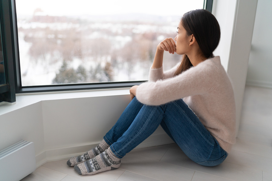 A woman sitting on the floor, gazing out of a window with a thoughtful expression on her face.