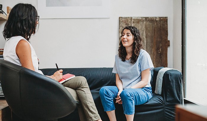 A female sits on a couch, smiling with her therapist, happy to give SAINT TMS therapy a chance for helping her depression.