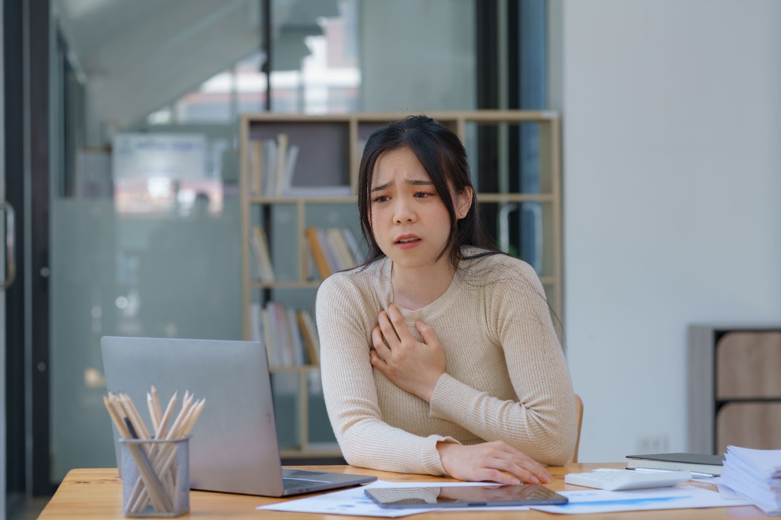 Asian woman with chest pain sitting at desk, experiencing panic disorder and panic attacks, considering TMS therapy.