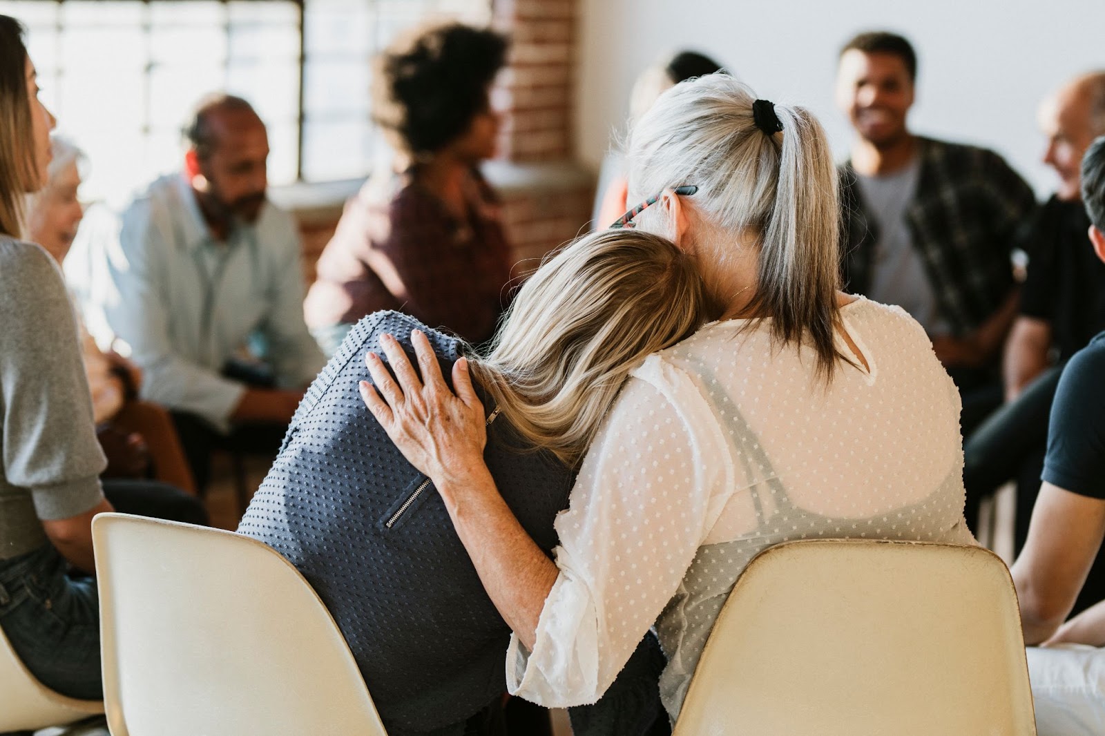 Two women sitting in chairs one hugging the other surrounded by a group of people. Keywords OCD anxiety TMS therapy.