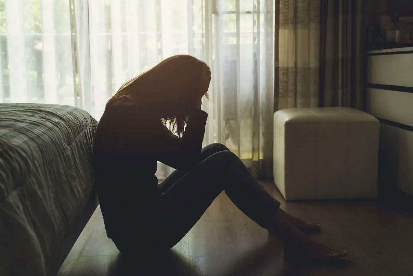 A woman sits on the floor in front of a bed, contemplating her thoughts in a serene bedroom setting.